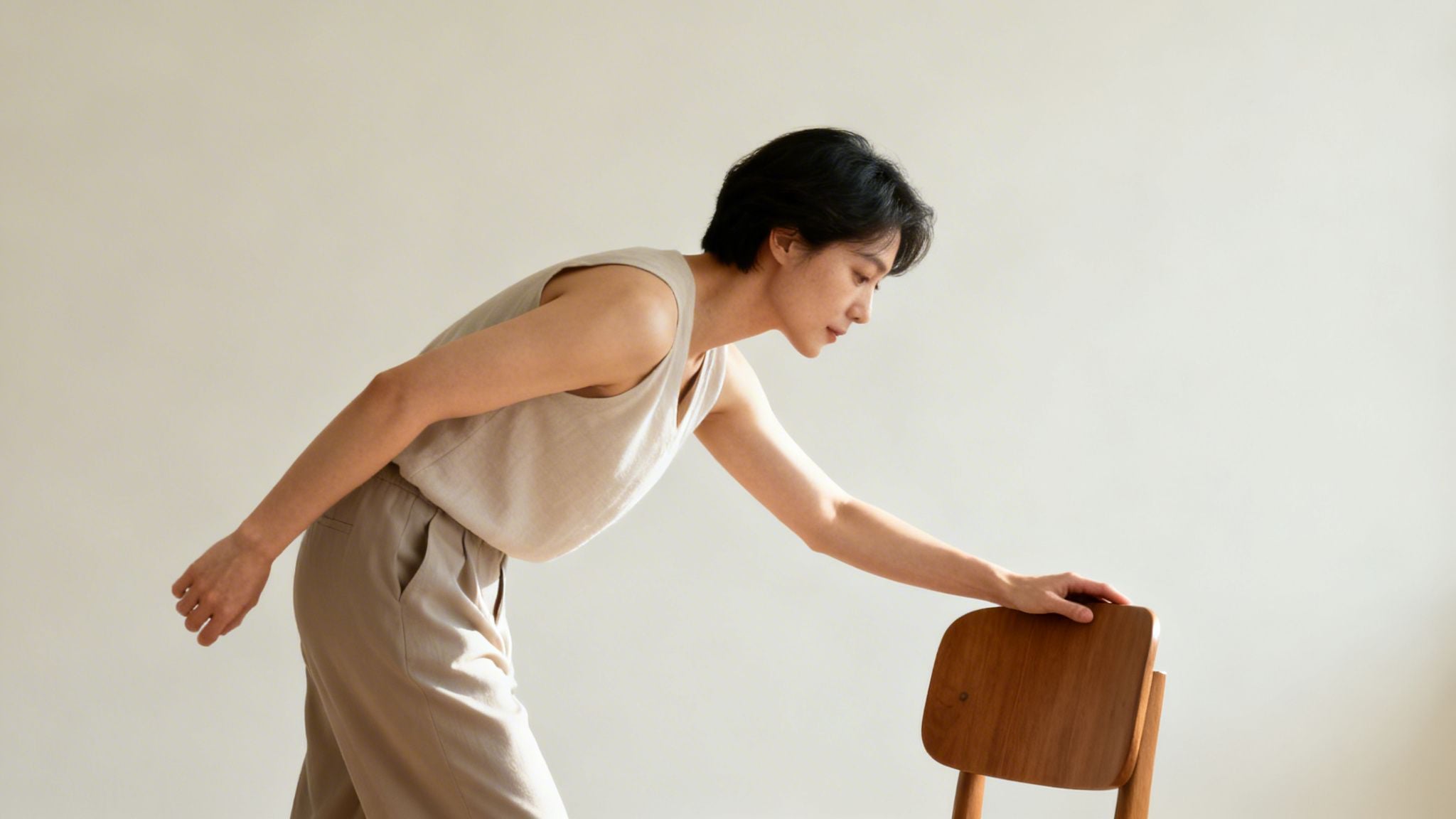 A woman in light clothing bends forward, reaching for a wooden chair, likely exercising.