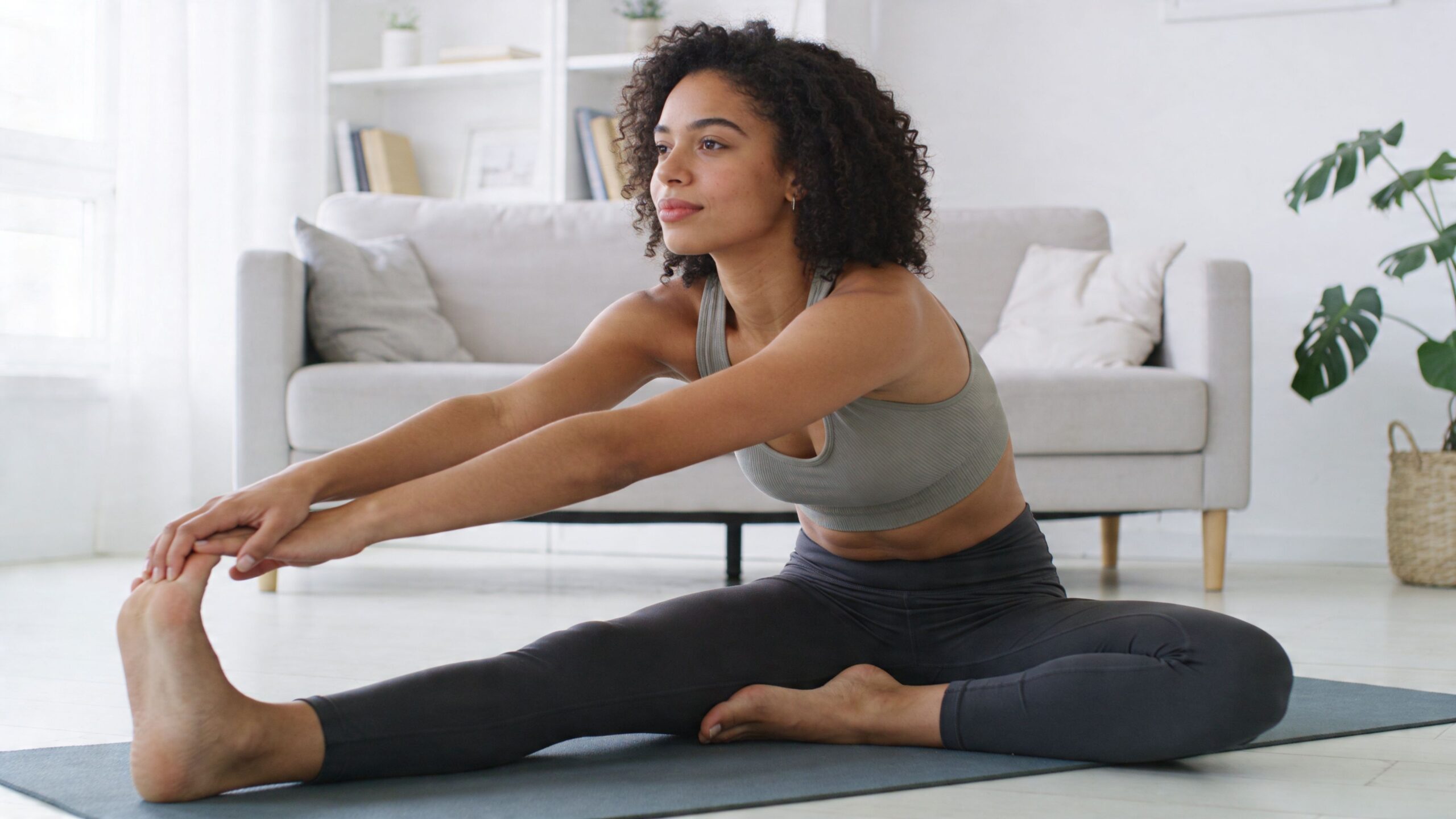 A fit woman with curly hair performs a seated hamstring stretch on a yoga mat in a living room.