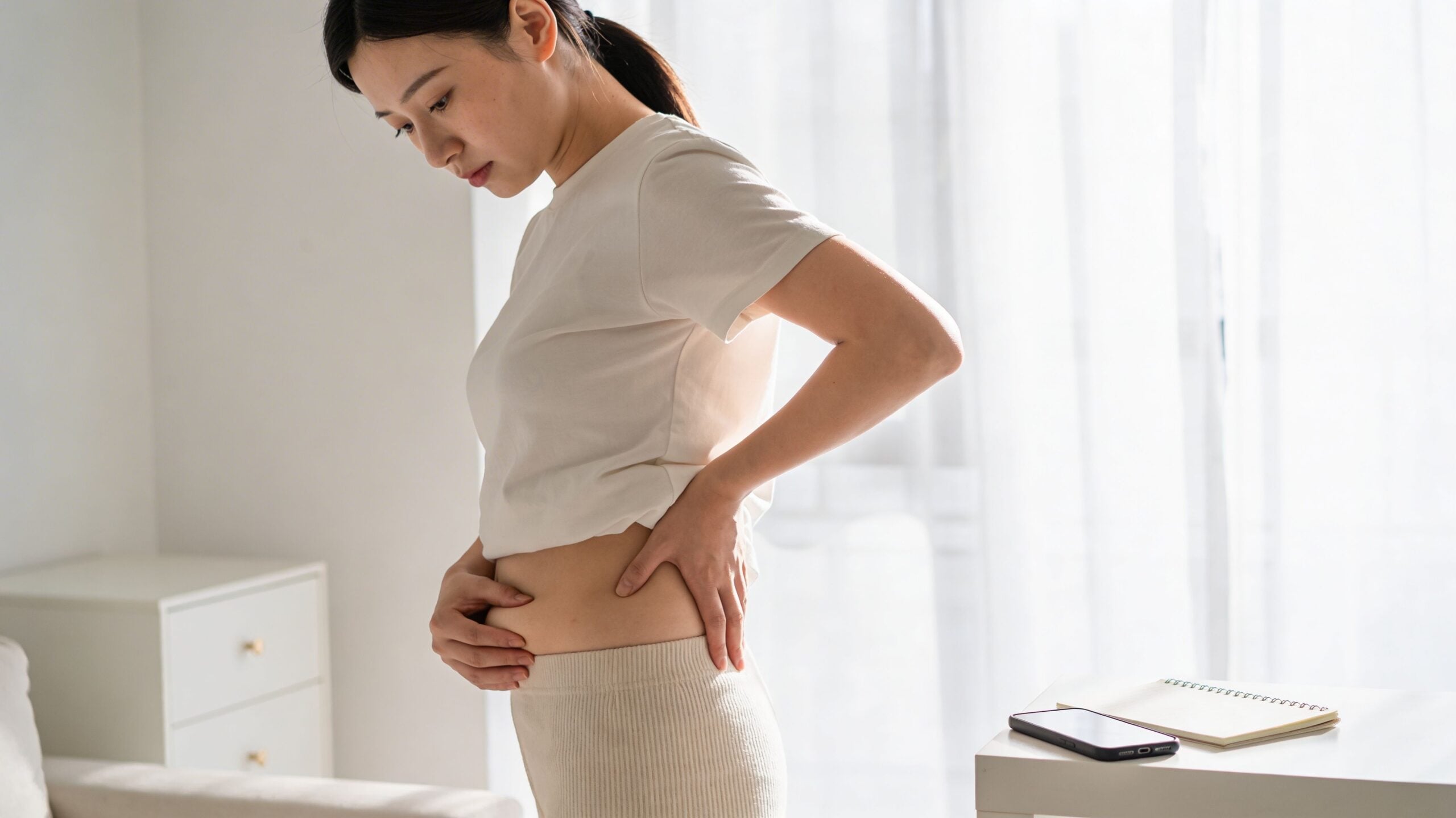 A woman standing sideways in her home touching her lower abdomen and hip area with both hands.