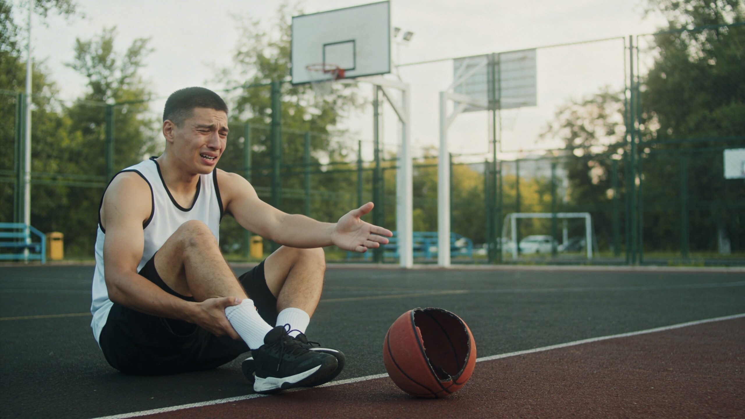 A young basketball player sitting on the court holding his injured ankle in pain during a game.