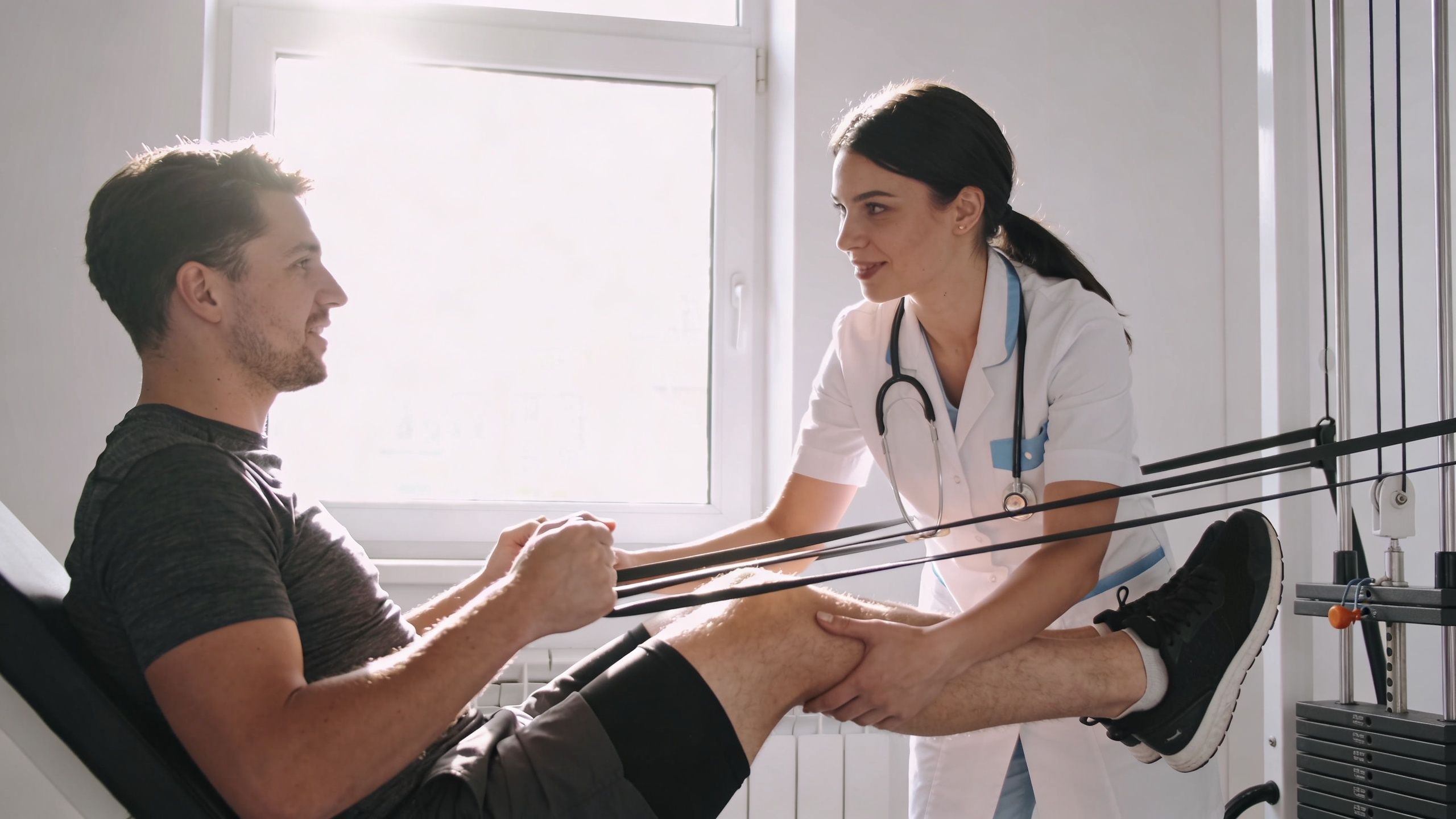A female physical therapist assisting a male patient with leg rehabilitation exercises using resistance bands in a clinic.