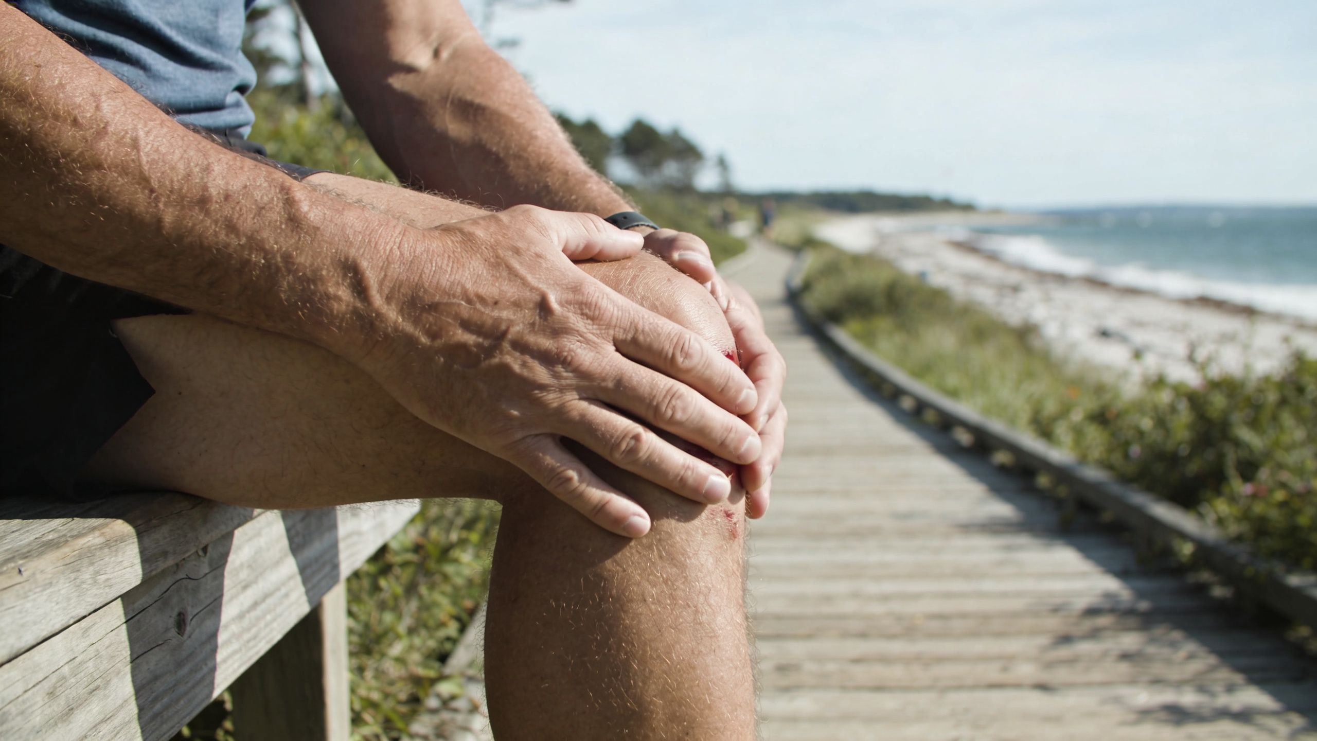 A person sitting on a wooden bench clutching their knee, which has a small open wound.