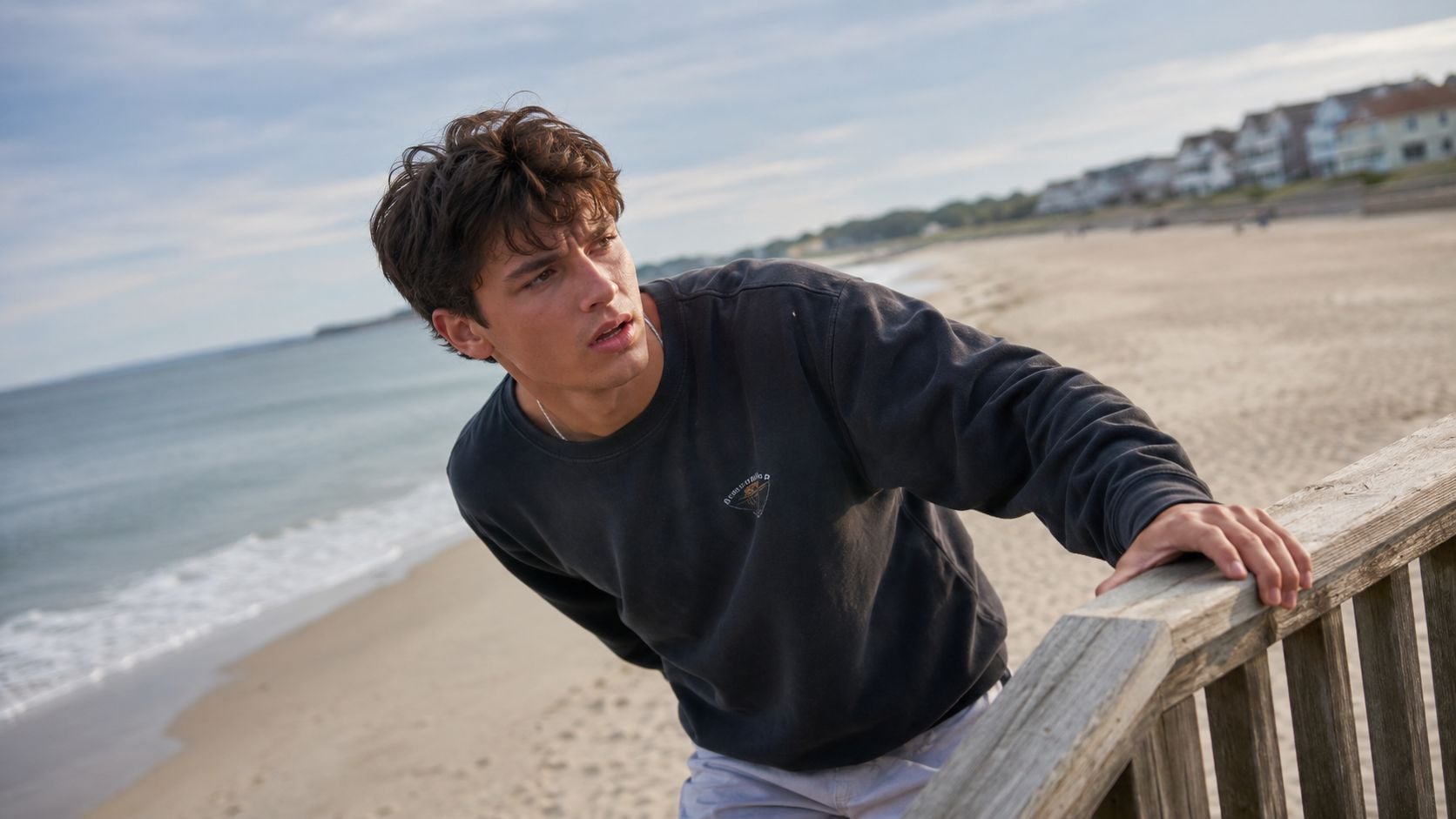 A young man looking concerned while leaning on a wooden railing on a beach near the ocean.
